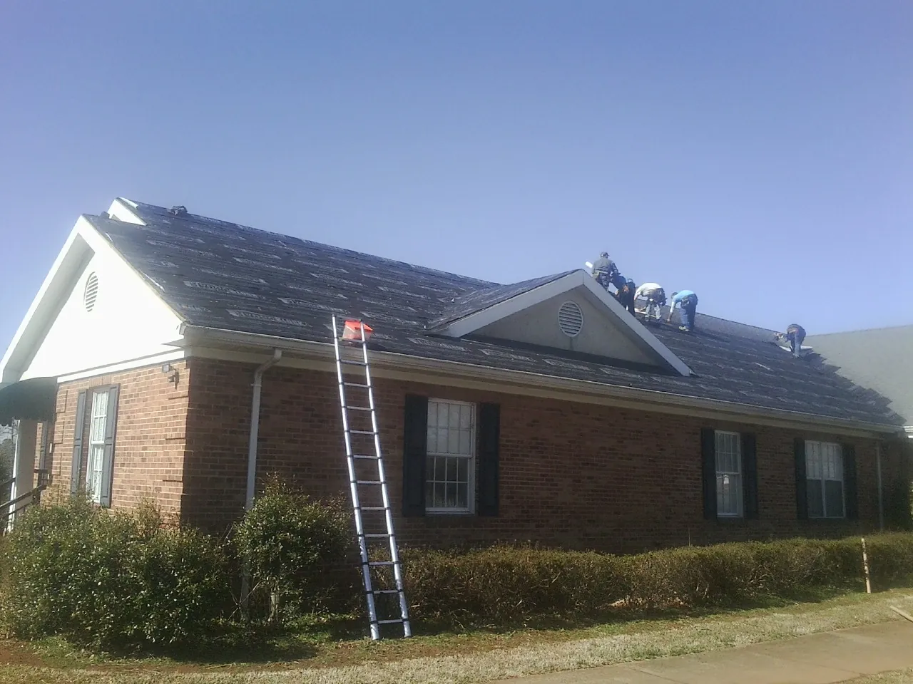 Crew tearing shingles off a brick ranch roof with a ladder set — Metal Roof Repair work by our crew in Royston