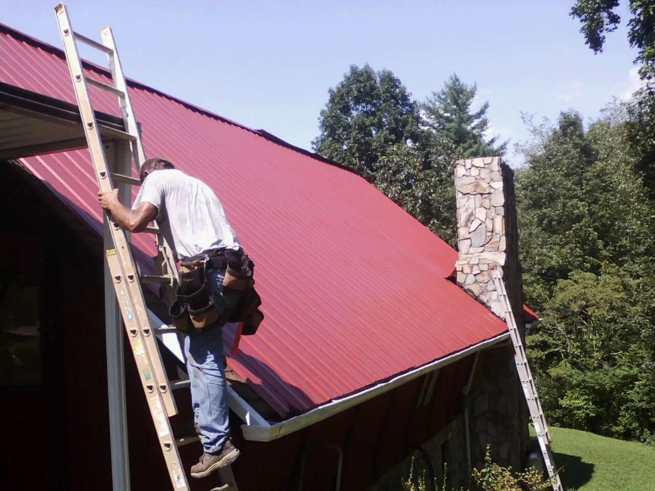 Roofer on a ladder beside a red metal roof and stone chimney — Soffit & Fascia Repair by Roofing Brothers in Royston